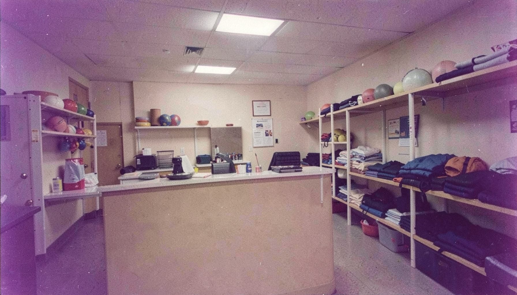Interior of a community recreation office with a service counter, shelves holding sports balls and folded equipment, soft fluorescent lighting, analog film grain