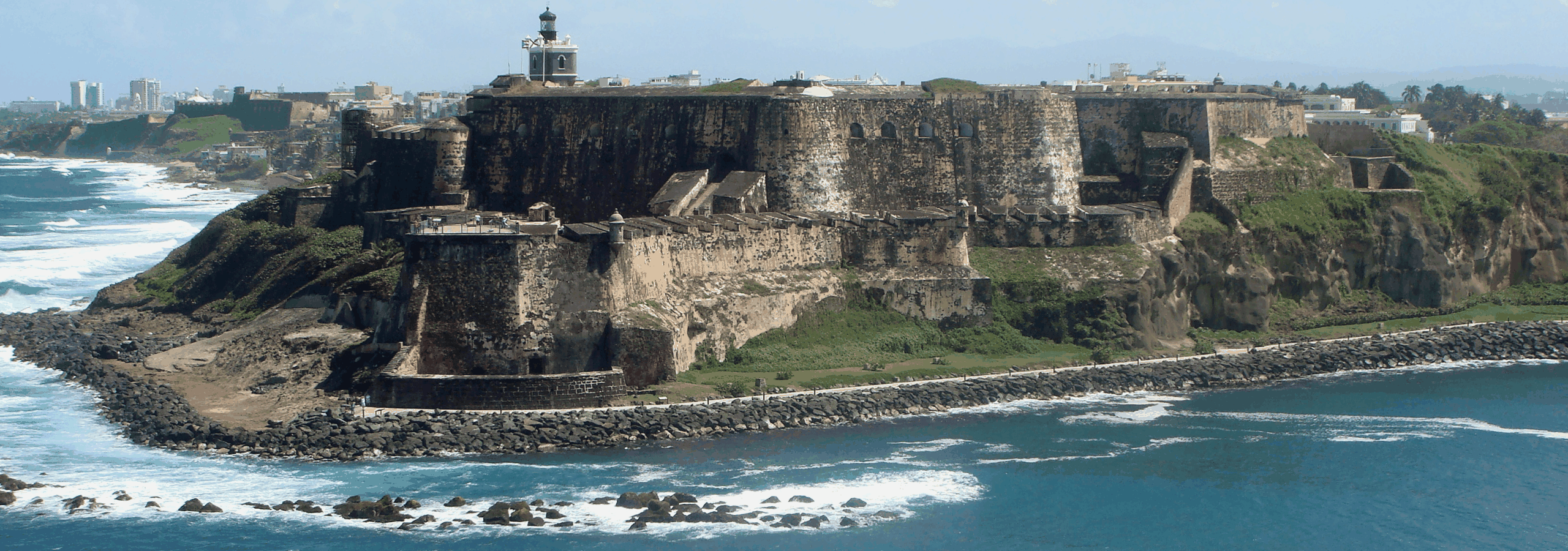 Aerial view of a large stone coastal fortress on a cliff, with ocean waves, a lighthouse tower, and a distant cityscape